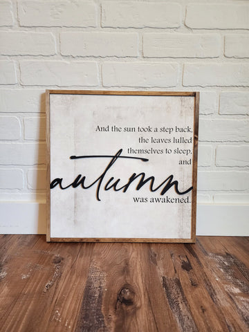 framed wood sign with the quote &quot;and the sun took a step back, the leaves lulled themselves to sleep, and autumn was awakened.&quot; The background is a beige/cream rustic looking texture, the writing is black, the frame is a warm brown stain.